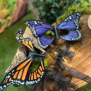 May include: Two butterfly masks with black frames and colourful wings. One mask has orange and black wings with white dots, the other has purple and black wings with white dots. Black ribbons are attached to the masks.