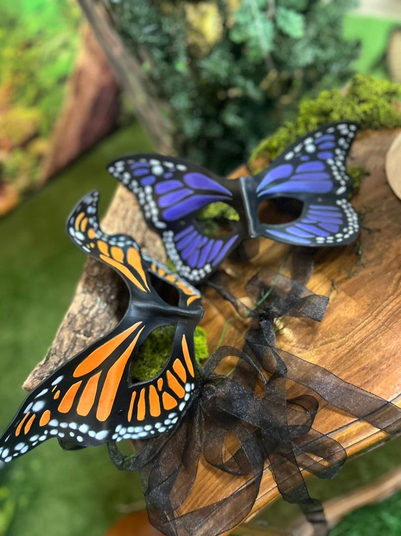 May include: Two butterfly masks with black frames and colourful wings. One mask has orange and black wings with white dots, the other has purple and black wings with white dots. Black ribbons are attached to the masks.