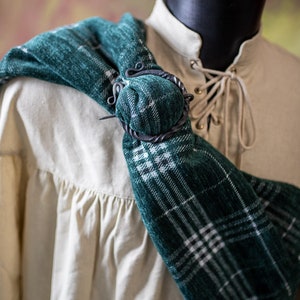 May include: A green and white plaid scarf with a black metal clasp. The scarf is draped over a white shirt with a lace-up collar.