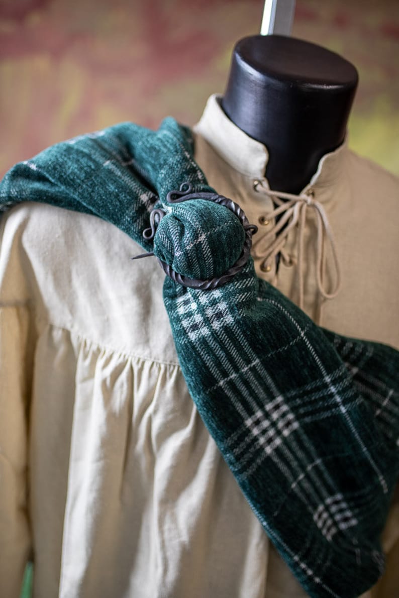 May include: A green and white plaid scarf with a black metal clasp. The scarf is draped over a white shirt with a lace-up collar.