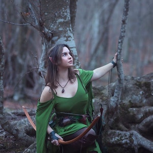 May include: A woman dressed as an elf in a green dress and brown boots holds a bow and arrow in a woodland setting.