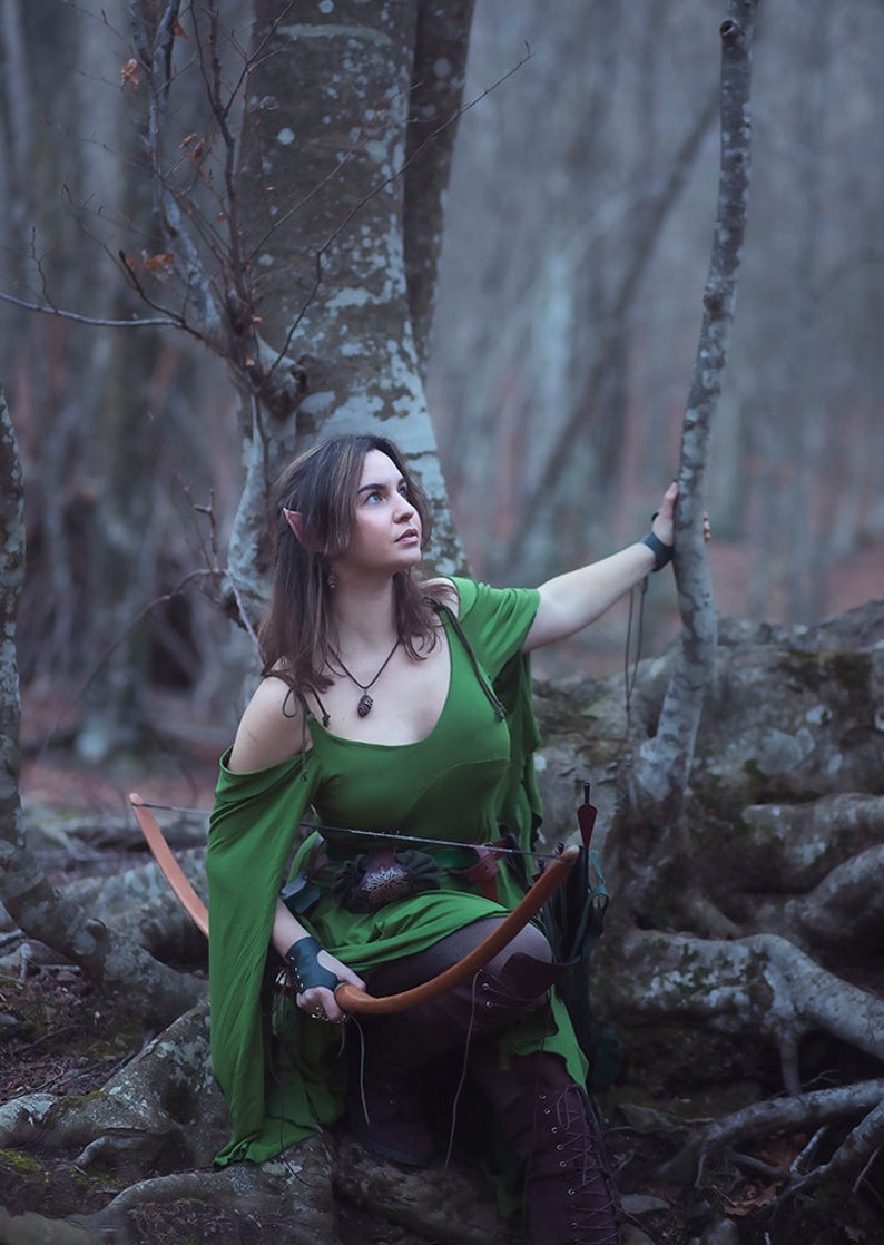 May include: A woman dressed as an elf in a green dress and brown boots holds a bow and arrow in a woodland setting.