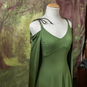 May include: A green, sleeveless dress with a V-neckline and a flowing skirt. The dress has a tie at the shoulder and is on a white mannequin.