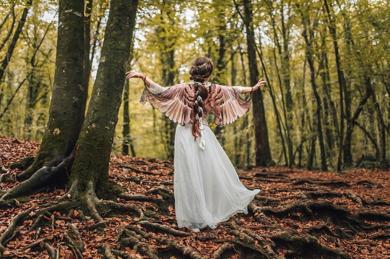 May include: A woman wearing a white dress and a brown and pink feathered wing shawl stands in a forest with fall leaves on the ground.