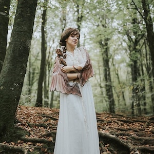 May include: A woman in a white dress and brown shawl stands in a forest with fallen leaves on the ground.