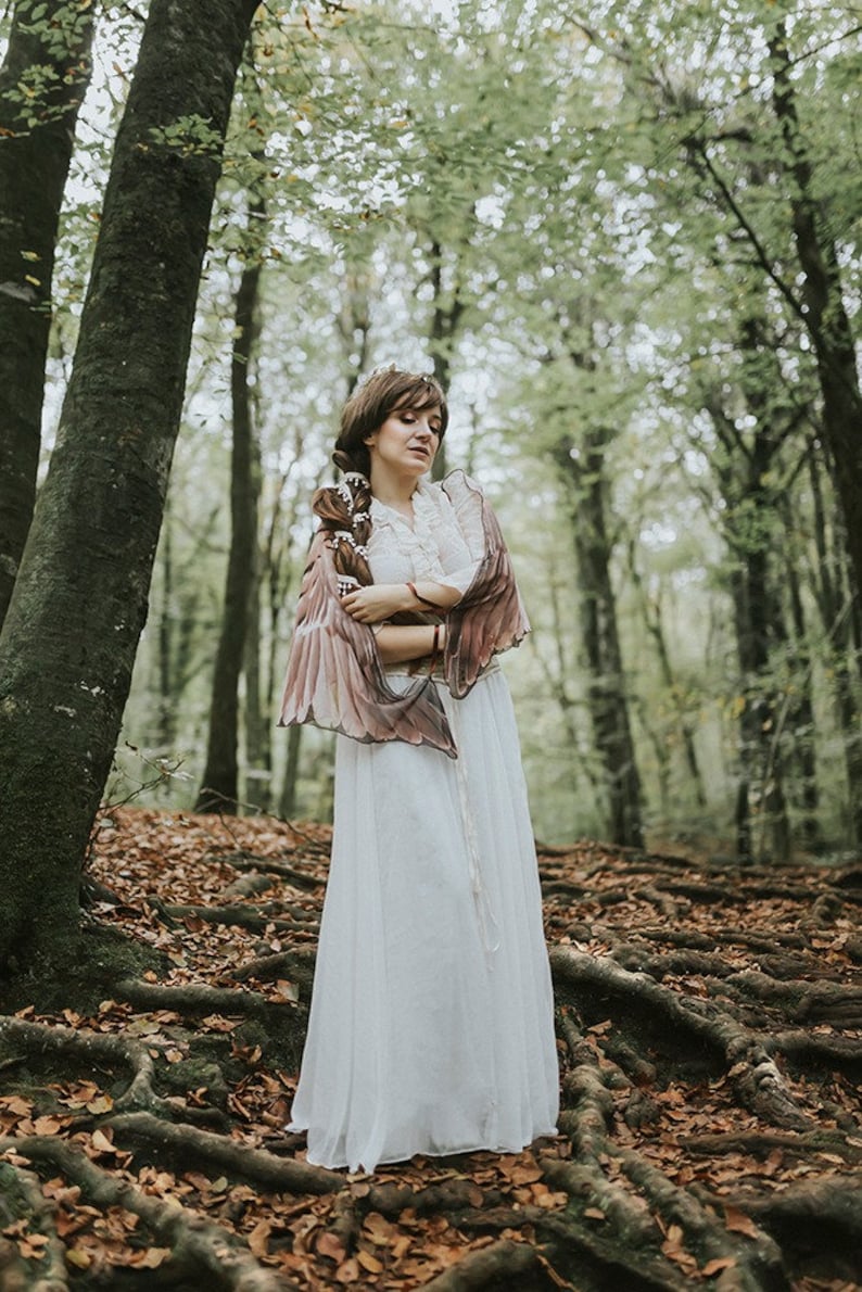 May include: A woman in a white dress and brown shawl stands in a forest with fallen leaves on the ground.