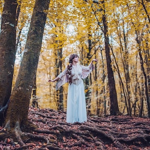 May include: A woman in a white dress with feathered sleeves stands in a forest with yellow leaves. She is looking up and her arms are outstretched.
