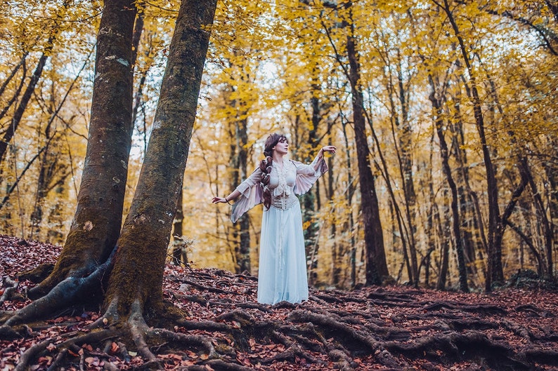 May include: A woman in a white dress with feathered sleeves stands in a forest with yellow leaves. She is looking up and her arms are outstretched.