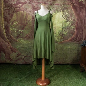 May include: A green sleeveless dress with a high low hemline and long flowing sleeves. The dress is on a wooden mannequin against a backdrop of a forest scene.