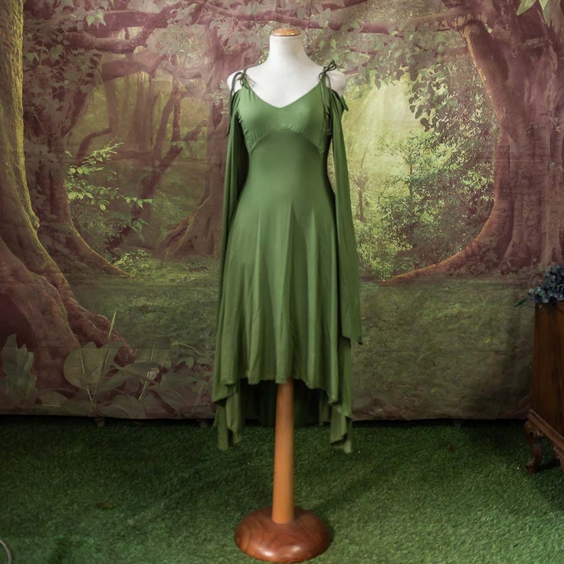 May include: A green sleeveless dress with a high low hemline and long flowing sleeves. The dress is on a wooden mannequin against a backdrop of a forest scene.