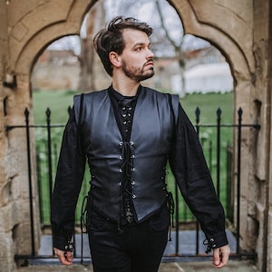 K&ouml;nnte beinhalten: Ein Mann tr&auml;gt eine schwarze Lederweste mit Schn&uuml;rung vorne, ein schwarzes Langarmhemd und eine schwarze Jeans. Er steht vor einem Steinbogen.