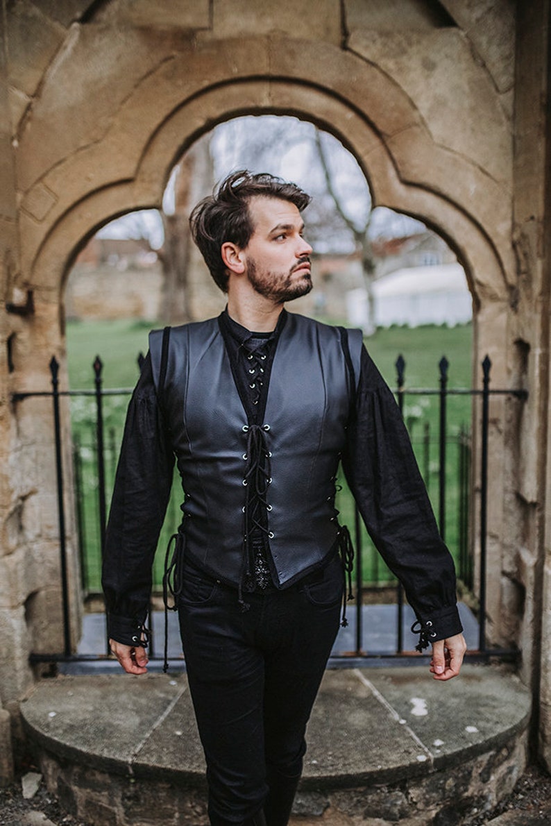 K&ouml;nnte beinhalten: Ein Mann tr&auml;gt eine schwarze Lederweste mit Schn&uuml;rung vorne, ein schwarzes Langarmhemd und eine schwarze Jeans. Er steht vor einem Steinbogen.