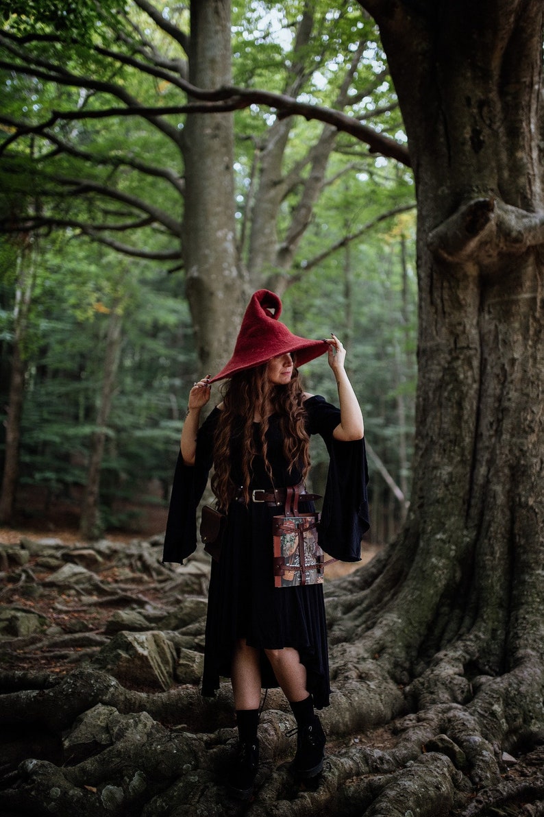 May include: A woman wearing a red pointed hat and a black dress with a leather belt and pouch stands in a forest.