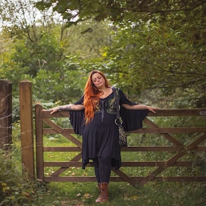 May include: A woman with long red hair stands by a wooden gate in a forest. She is wearing a black dress with a green and black patterned vest and brown boots.