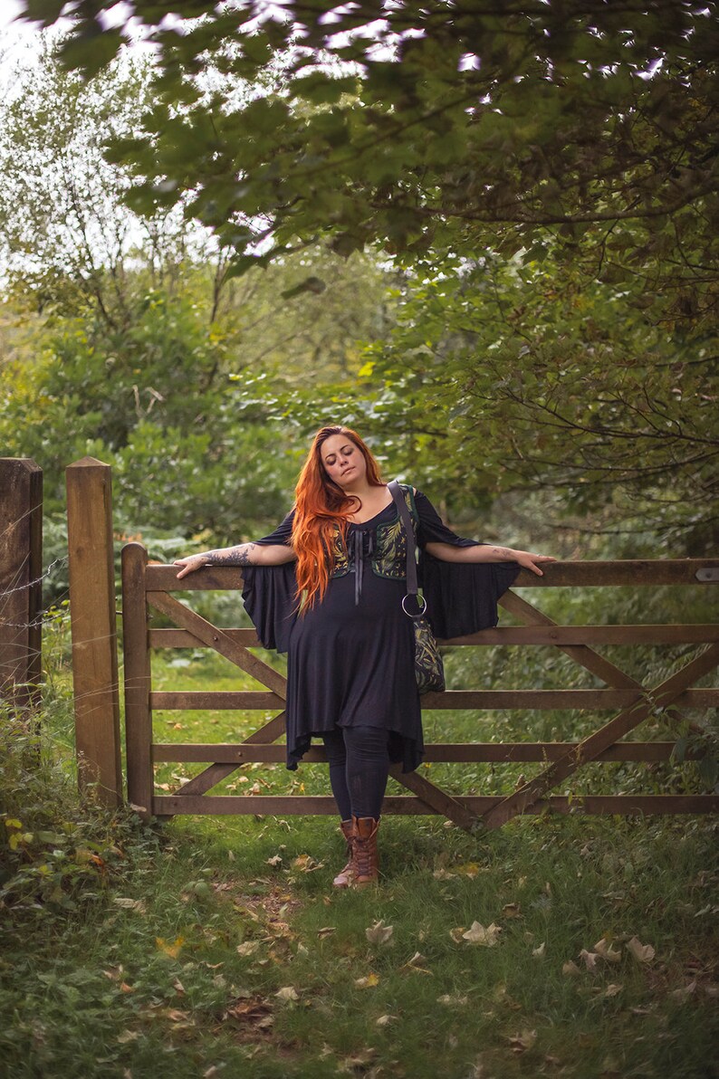 May include: A woman with long red hair stands by a wooden gate in a forest. She is wearing a black dress with a green and black patterned vest and brown boots.