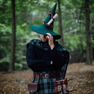 May include: A person wearing a green pointed hat with a rolled scroll attached to the side, a black shirt, a green and black plaid kilt, and a brown leather belt with a pouch and a book attached.