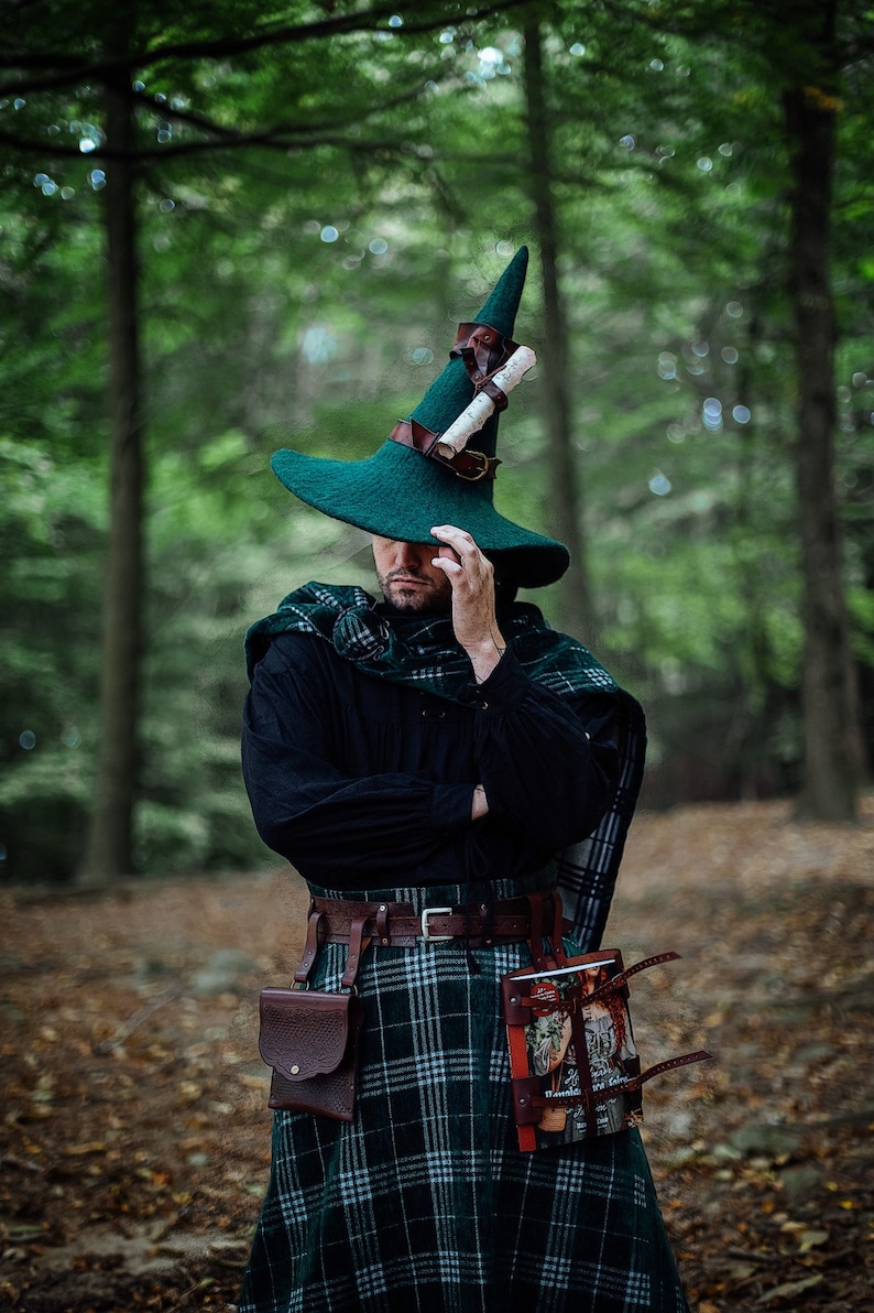 May include: A person wearing a green pointed hat with a rolled scroll attached to the side, a black shirt, a green and black plaid kilt, and a brown leather belt with a pouch and a book attached.