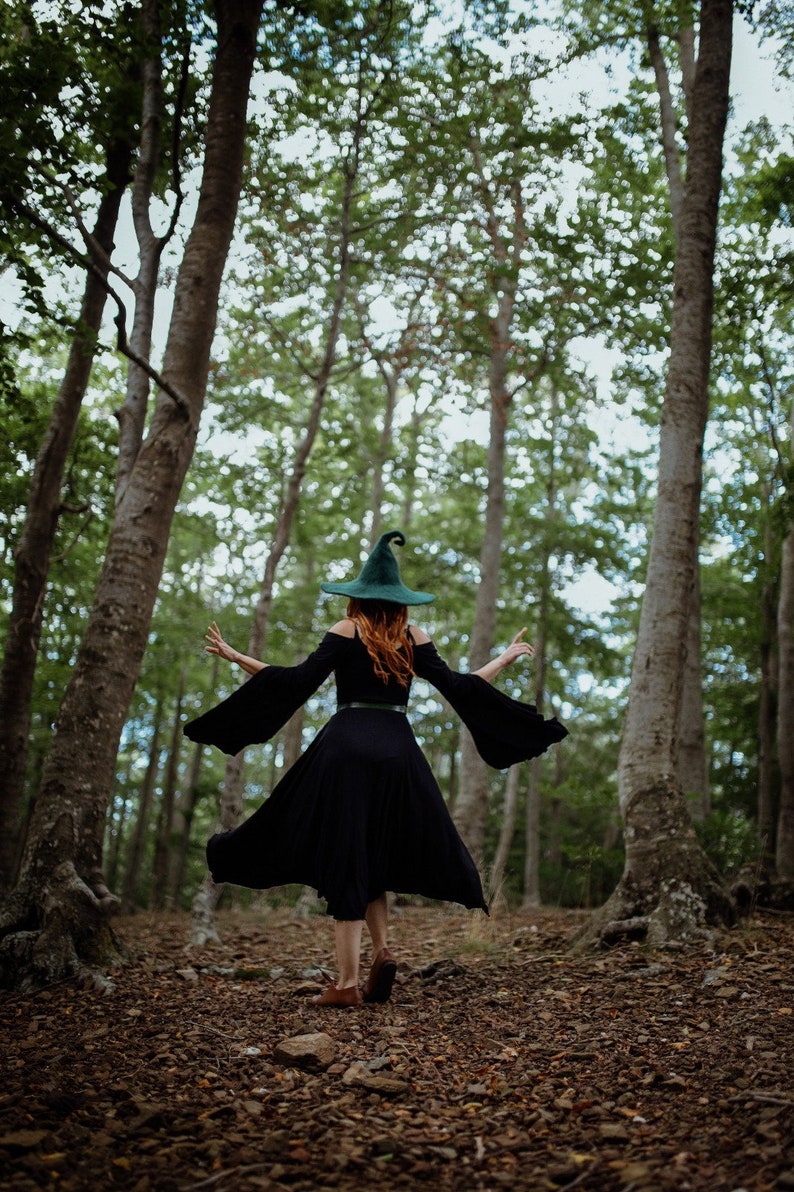 May include: A woman wearing a black dress and a green pointed hat walks through a forest. The forest floor is covered in fallen leaves.