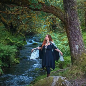 May include: A woman wearing a black dress and a large, white butterfly wing costume stands on a mossy rock by a stream in a forest. The stream is surrounded by lush green foliage.