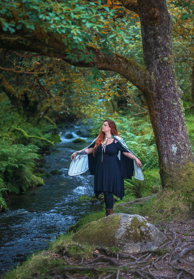May include: A woman wearing a black dress and a large, white butterfly wing costume stands on a mossy rock by a stream in a forest. The stream is surrounded by lush green foliage.