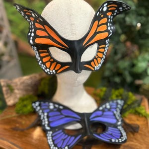 May include: Two butterfly masks. The top mask is black with orange and white details, resembling a monarch butterfly. The bottom mask is purple with white details. Both masks are on a wooden surface.