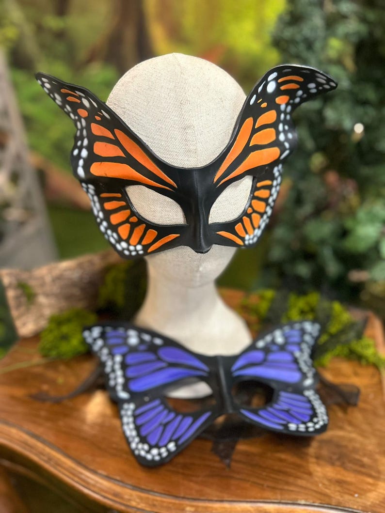 May include: Two butterfly masks. The top mask is black with orange and white details, resembling a monarch butterfly. The bottom mask is purple with white details. Both masks are on a wooden surface.