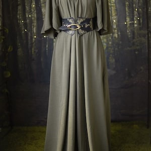 K&ouml;nnte beinhalten: Ein langes, olivgr&uuml;nes Kleid mit einem breiten, schwarzen G&uuml;rtel mit einer goldenen Schnalle und einem verzierten Design. Das Kleid hat einen leicht schulterfreien Ausschnitt und kurze, weite &Auml;rmel.