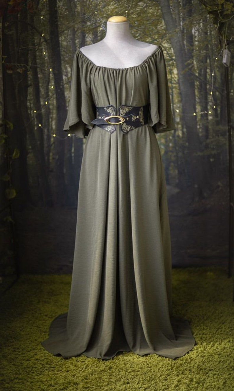 K&ouml;nnte beinhalten: Ein langes, olivgr&uuml;nes Kleid mit einem breiten, schwarzen G&uuml;rtel mit einer goldenen Schnalle und einem verzierten Design. Das Kleid hat einen leicht schulterfreien Ausschnitt und kurze, weite &Auml;rmel.