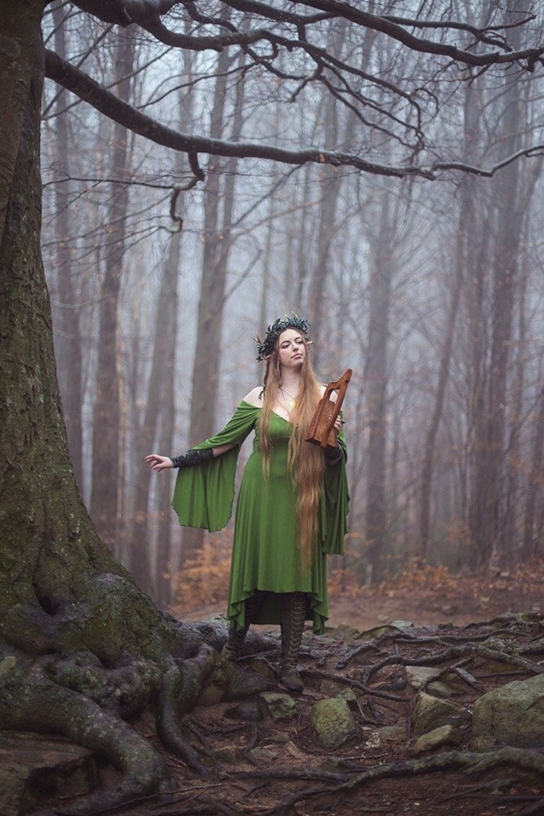 May include: A woman wearing a green dress and a leaf crown stands in a misty woodland. She holds a wooden lyre in her right hand. The woodland is filled with bare trees and fallen leaves.