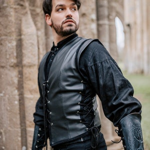 K&ouml;nnte beinhalten: Ein Mann tr&auml;gt eine schwarze Lederweste mit Schn&uuml;rung vorne und eine schwarze Hose. Er tr&auml;gt schwarze Handschuhe und ein schwarzes Hemd mit hohem Kragen. Die Weste hat ein dekoratives Design auf der R&uuml;ckseite.