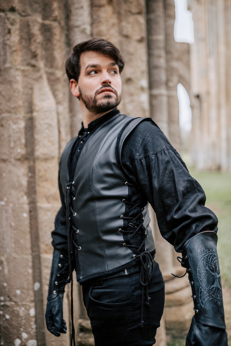 K&ouml;nnte beinhalten: Ein Mann tr&auml;gt eine schwarze Lederweste mit Schn&uuml;rung vorne und eine schwarze Hose. Er tr&auml;gt schwarze Handschuhe und ein schwarzes Hemd mit hohem Kragen. Die Weste hat ein dekoratives Design auf der R&uuml;ckseite.
