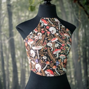 May include: A black halter top with a mushroom and fern print. The print features red, white, and brown mushrooms, green ferns, and other forest elements on a black background.