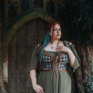 K&ouml;nnte beinhalten: Eine Frau mit roten und blauen Haaren tr&auml;gt ein gr&uuml;nes Kleid mit einem karierten Korsett und braunen Lederakzenten. Sie steht vor einer Holzt&uuml;r mit einem gew&ouml;lbten Steinrahmen.