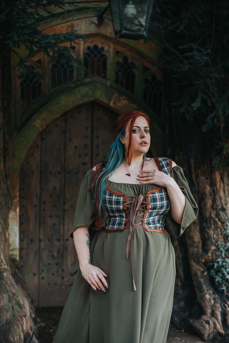 K&ouml;nnte beinhalten: Eine Frau mit roten und blauen Haaren tr&auml;gt ein gr&uuml;nes Kleid mit einem karierten Korsett und braunen Lederakzenten. Sie steht vor einer Holzt&uuml;r mit einem gew&ouml;lbten Steinrahmen.
