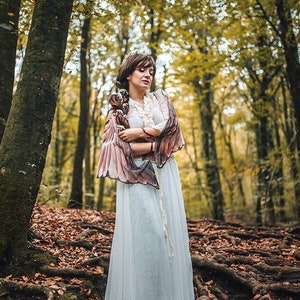 May include: A woman wearing a white dress and a brown and pink feathered shawl stands in a forest with fallen leaves on the ground.