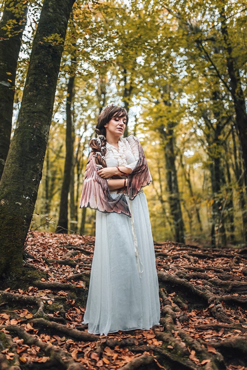 May include: A woman wearing a white dress and a brown and pink feathered shawl stands in a forest with fallen leaves on the ground.
