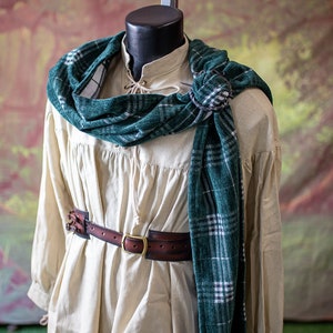 May include: A cream-colored shirt with long sleeves and a gathered waist. A brown leather belt with a brass buckle is worn over the shirt. A green and white plaid scarf is draped around the neck and tied at the shoulder.