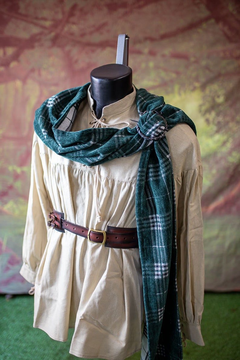 May include: A cream-colored shirt with long sleeves and a gathered waist. A brown leather belt with a brass buckle is worn over the shirt. A green and white plaid scarf is draped around the neck and tied at the shoulder.