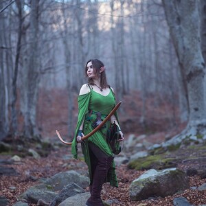 May include: A woman dressed as an elf in a green dress and brown boots holds a bow and arrow in a woodland setting.