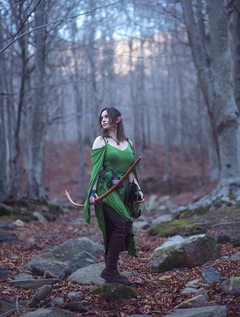May include: A woman dressed as an elf in a green dress and brown boots holds a bow and arrow in a woodland setting.