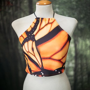 May include: A halter top with a butterfly print in shades of orange, black, and white. The top has a thin black strap that ties at the neck.