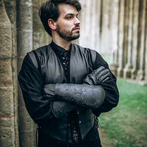 K&ouml;nnte beinhalten: Ein Mann tr&auml;gt eine schwarze Lederweste mit Schn&uuml;rung vorne, ein schwarzes Hemd und schwarze Lederarmsch&uuml;tzer mit aufwendigen Designs. Er steht vor einer Steinmauer.