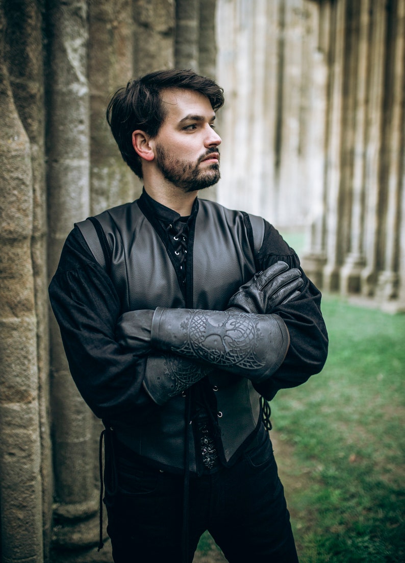 K&ouml;nnte beinhalten: Ein Mann tr&auml;gt eine schwarze Lederweste mit Schn&uuml;rung vorne, ein schwarzes Hemd und schwarze Lederarmsch&uuml;tzer mit aufwendigen Designs. Er steht vor einer Steinmauer.