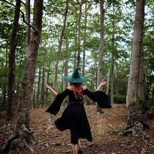 May include: A woman wearing a green pointed witch hat and a black dress walks through a forest.