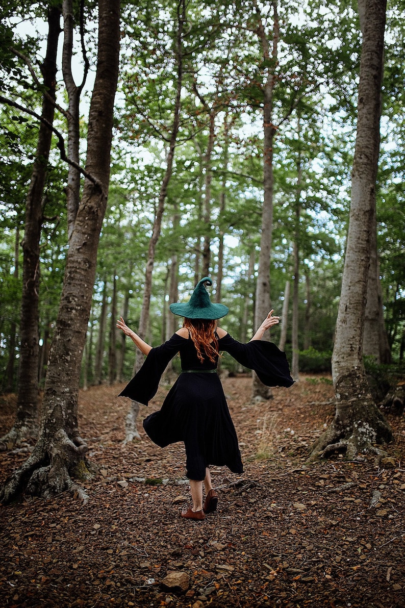 May include: A woman wearing a green pointed witch hat and a black dress walks through a forest.