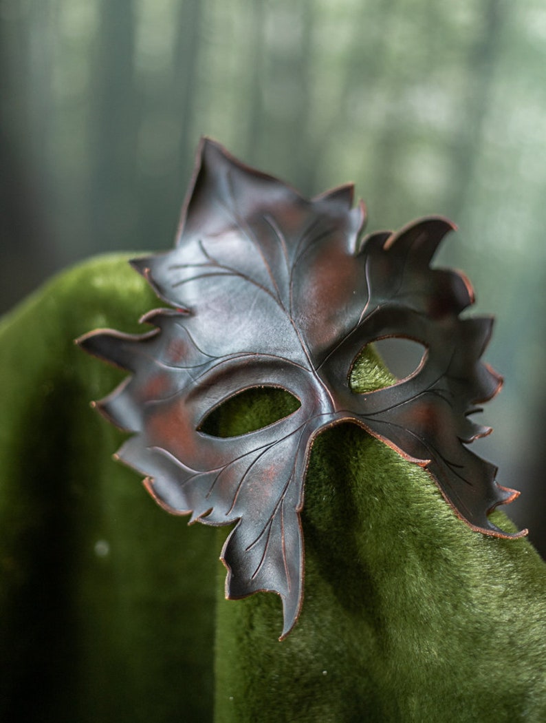 Puede incluir: Una m&aacute;scara de cuero marr&oacute;n oscuro con forma de hoja con detalles intrincados. La m&aacute;scara tiene agujeros para los ojos y una punta puntiaguda.