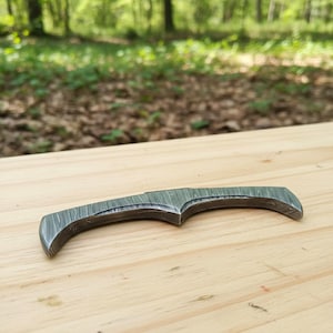 May include: A small, curved knife with a patterned, textured blade. The knife is made of metal and has a dark, gray color. The knife is resting on a light-colored wooden surface.