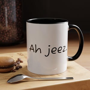 May include: White ceramic mug with a black interior and handle. The mug features the phrase "Ah jeez" in black lettering. A spoon, a macaron, and coffee beans are on a wooden surface.