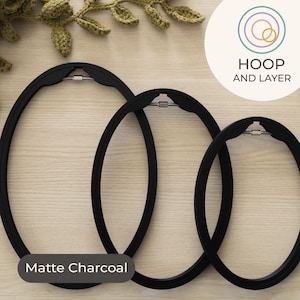 Puede incluir: Tres bastidores de bordado ovalados en negro mate de diferentes tamaños se muestran sobre una superficie de madera clara. Los bastidores tienen un diseño moderno con un cierre metálico. El texto "Matte Charcoal" es visible. La imagen también incluye el texto "HOOP AND LAYER".