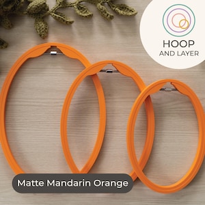 May include: Three matte mandarin orange oval embroidery hoops of varying sizes are displayed on a light wood surface. The hoops are made of a smooth material and have a silver metal clasp. The text "Matte Mandarin Orange" is displayed on the bottom of the image.